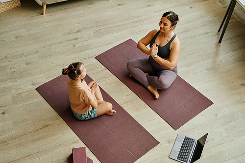 High angle view at female instructor assisting girl with down syndrome doing yoga and sitting in lotus position on mats