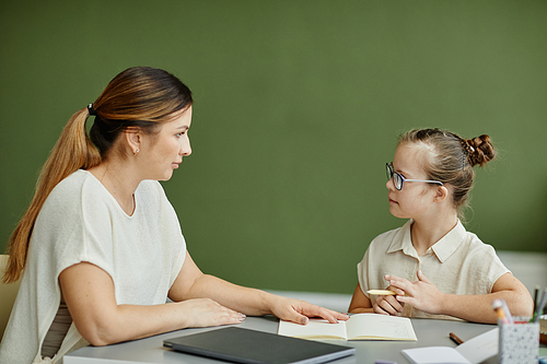 Minimal side view portrait of young mother or female tutor homeschooling girl with down syndrome against green wall