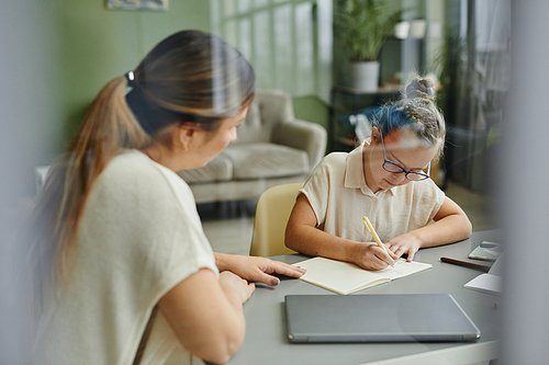 Portrait of young girl with down syndrome studying at home with female tutor helping, behind glass view