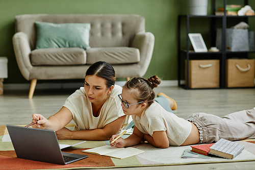 Portrait of young mother helping girl with down syndrome studying at home while lying on floor together, non traditional education