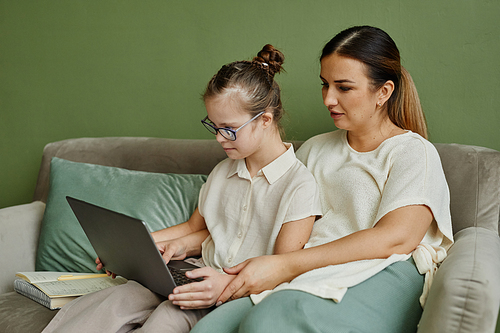 Portrait of mother and daughter with down syndrome relaxing on couch together in cozy home and using laptop, copy space