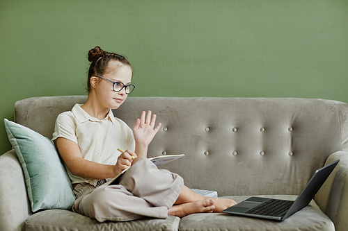 Minimal portrait of young girl with down syndrome studying online at home while sitting on cozy couch and waving to camera