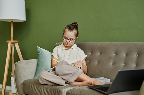 Minimal portrait of young girl with down syndrome studying at home while sitting on cozy couch against green wall, copy space