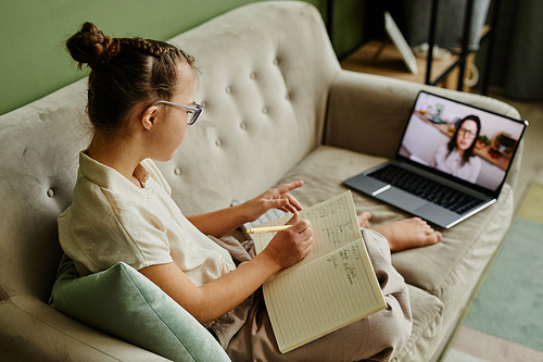 High angle portrait of young girl with down syndrome studying online at home while sitting on cozy couch and listening to tutor on screen