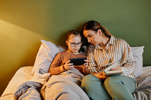 Minimal portrait of caring mother and daughter with down syndrome using tablet to watch cartoons at bedtime, copy space