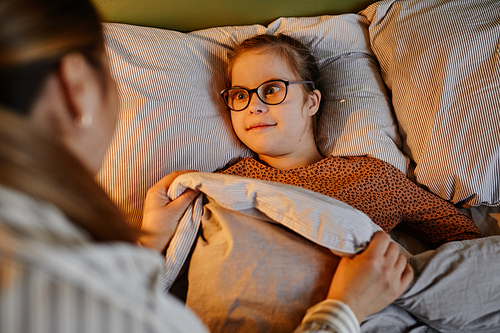 High angle portrait of caring mother tucking in child with down syndrome at bedtime in cozy lamp light, copy space