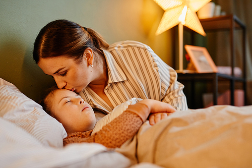 Portrait of caring mother kissing child with down syndrome at bedtime lit by cozy lamp light, copy space