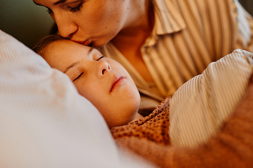 Close up of caring mother kissing child with down syndrome at bedtime lit by cozy lamp light, copy space