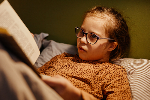 Portrait of young girl with down syndrome reading book in bed at night lit by cozy lamplight