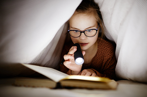 Front view portrait of young girl with down syndrome reading book under covers using flashlight, copy space