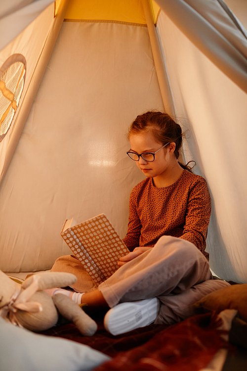 Vertical portrait of young girl with down syndrome reading book in cozy play tent lit by warm light