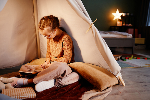 Portrait of young girl with down syndrome using tablet in cozy play tent at night, copy space