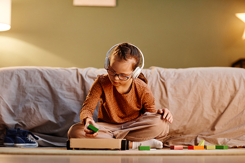 Portrait of young girl with down syndrome playing with toy blocks alone and wearing noise cancelling earphones, overstimulation concept, copy space