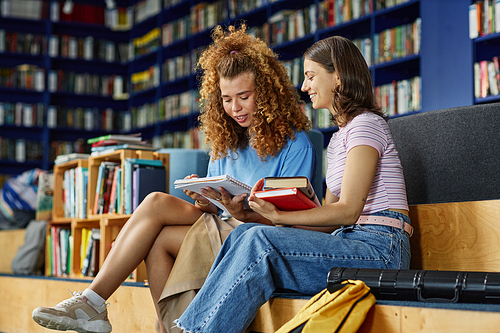 Vibrant portrait of two young girls reading books in college library and smiling, copy space