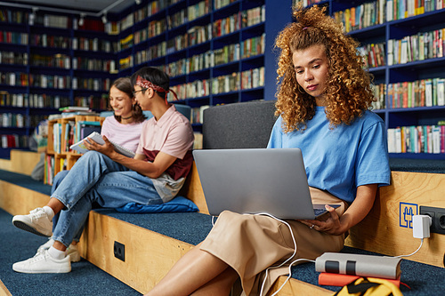 Portrait of curly haired young woman using laptop in college library and studying with people in background