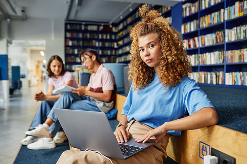 Portrait of curly haired young woman using laptop in college library and looking at camera with people in background, copy space