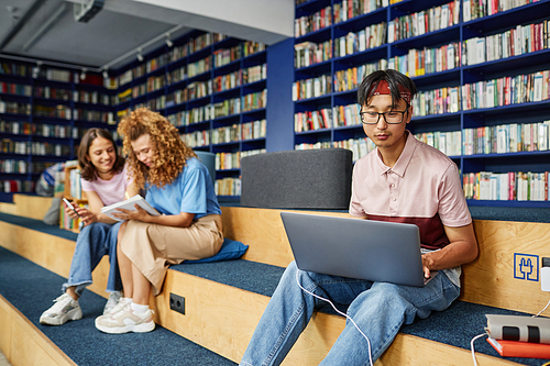 Vibrant portrait of Asian young man using laptop in college library and studying with people in background, copy space