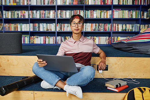 Vibrant full length portrait of young Asian man sitting cross legged with laptop in college library and looking at camera