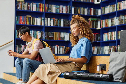 Side view portrait of curly haired young woman using laptop in library and smiling