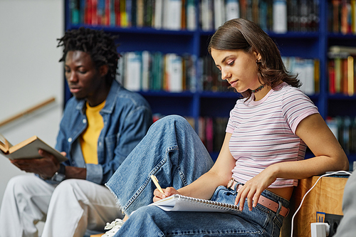 Vibrant side view portrait of young woman doing homework in college library and writing in notebook