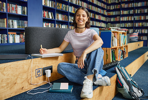 Vibrant full length portrait of smiling female student sitting cross legged in school library, copy space