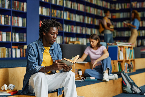 Portrait of young black man reading book in library lounge with students in background, copy space