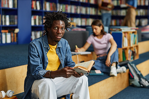 Vibrant portrait of young black man reading book in college library and smiling at camera, copy space