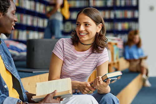 Vibrant shot of two young students chatting in college library and holding books