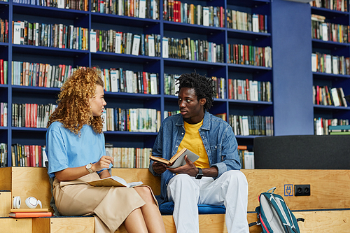 Portrait of two college students chatting in vibrant library setting with blue tone, copy space