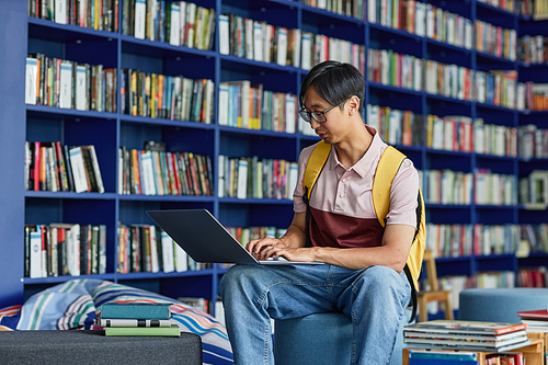 Portrait of young Asian man using laptop in library lounge with vibrant blue tones, copy space