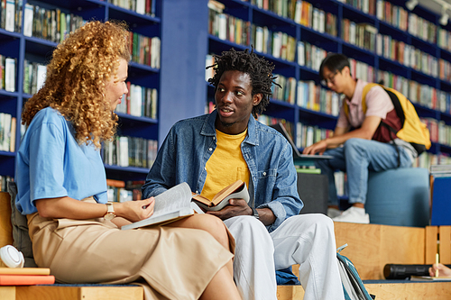 Diverse group of students studying in library lounge, focus on black young man talking to female friend