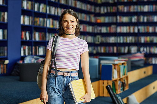 Vibrant waist up portrait of smiling young woman with backpack in college library looking at camera, copy space