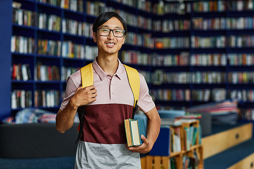 Waist up portrait of Asian young man with backpack standing in library and looking at camera smiling, copy space