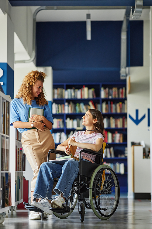 Vertical full length portrait of smiling young woman with disability talking to friend in college library