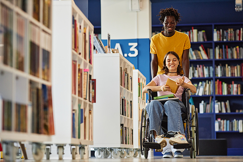 Vibrant full length shot of black young man assisting female student with disability in library and pushing wheelchair, copy space