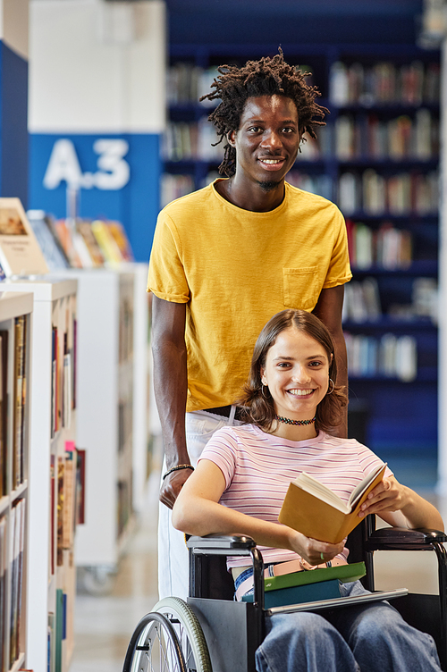 Vertical portrait of smiling young woman with disability looking at camera in college library with friend assisting