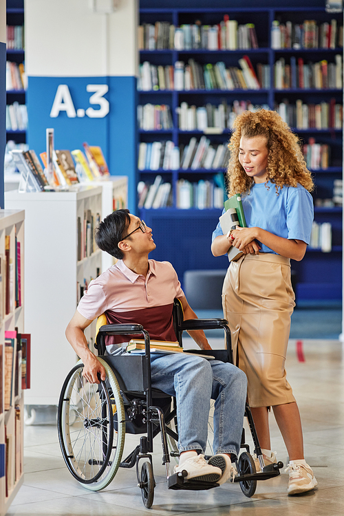 Vertical full length portrait of Asian student with disability talking to friend in college library setting