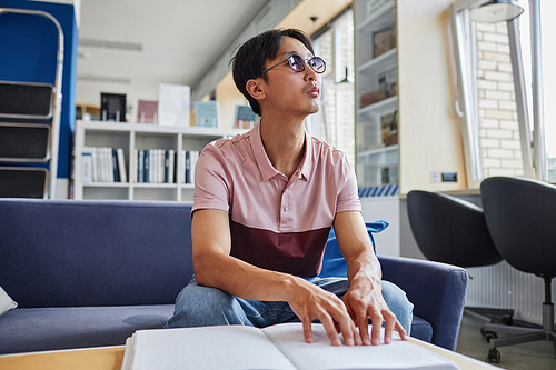 Low angle portrait of Asian young man reading book in tactile braille font in college library, copy space