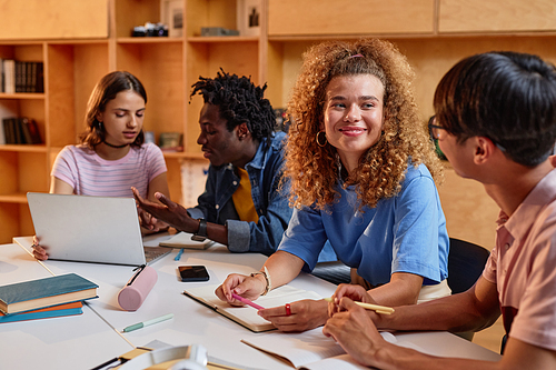 Diverse group of students working at table in library together, focus on smiling young woman with curly hair