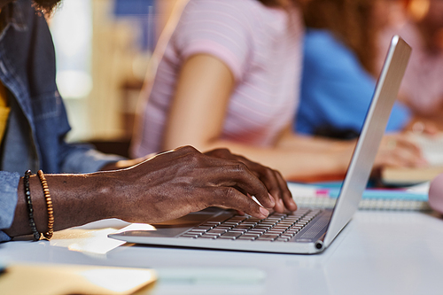 Close up of black young man typing on laptop keyboard while studying in college library with students in row, copy space