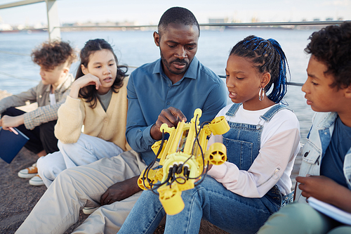 Portrait of young black girl holding robot model during engineering class outdoors with male teacher helping