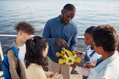 Portrait of black male teacher demonstrating robot model to group of children standing in circle during engineering class outdoors