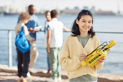 Waist up portrait of Asian teenage girl holding robot model and smiling at camera during outdoor engineering class with kids in background, copy space