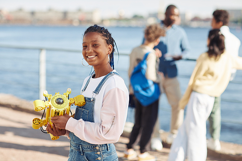 Waist up portrait of black teenage girl holding robot model and smiling at camera during outdoor engineering class with kids in background, copy space