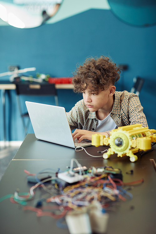 Vertical portrait of young curly haired boy using laptop in school during engineering class and programming robot