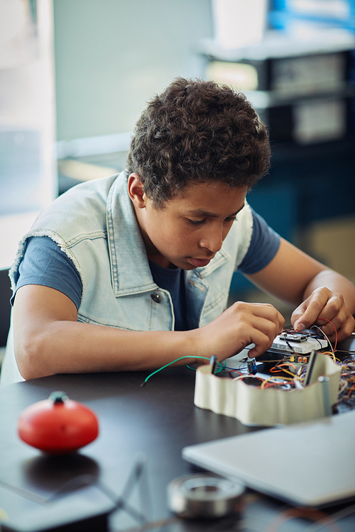Vertical portrait of black teenage boy building robot and wiring circuit panel during engineering class at school