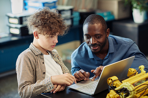 Portrait of black male teacher helping young boy building robot during engineering class in school and using laptop for programming together