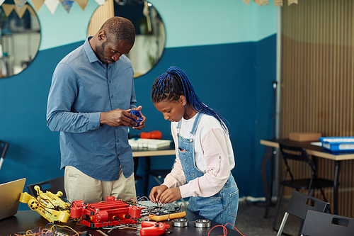 Portrait of male teacher helping young African American girl building robot during engineering class in school, copy space