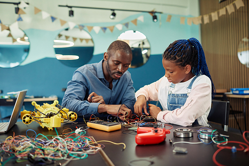 Portrait of black young girl building robots with male teacher helping during engineering class at school