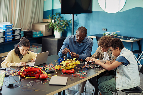 Portrait of black male teacher building robots with diverse group of children in engineering class at modern school, copy space